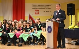 na zdjęciu widać przemawiającego dyrektora szkoły i siedzący w tle ch&oacute;r szkolny