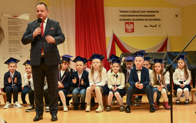 na zdjęciu widać przemawiającego dyrektora szkoły i siedzących pierwszoklasist&oacute;w