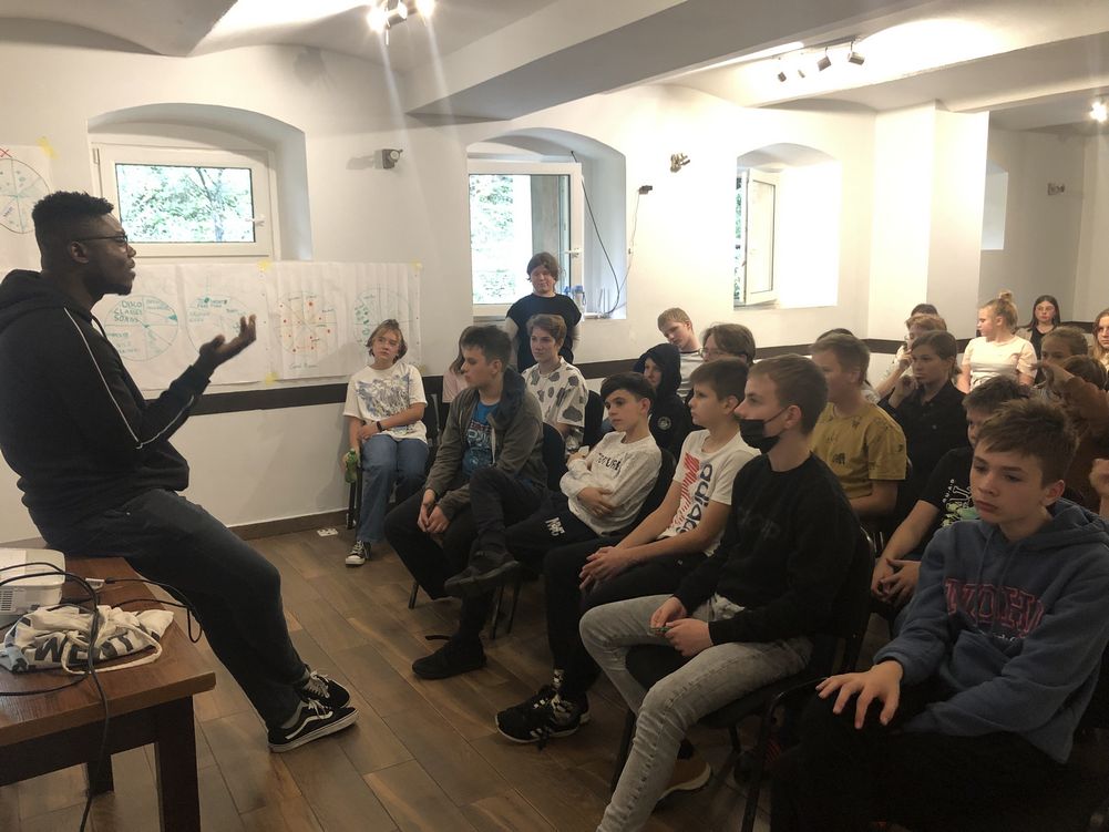 Na obrazku widać uczniów siedzących w sali i słuchających wykładu wolontariusza z Ghany.