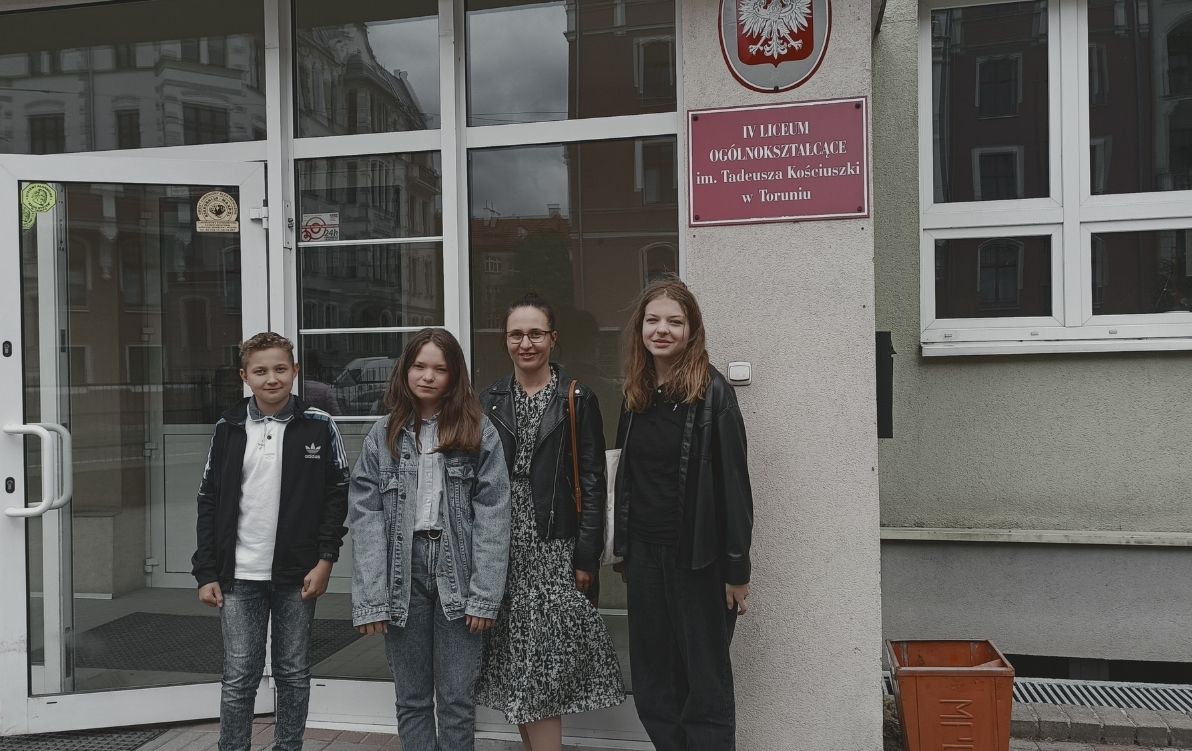 Liga Zadaniowa 2022 Na zdjęciu widać 3 uczniów z nauczycielką stojących przed budynkiem liceum