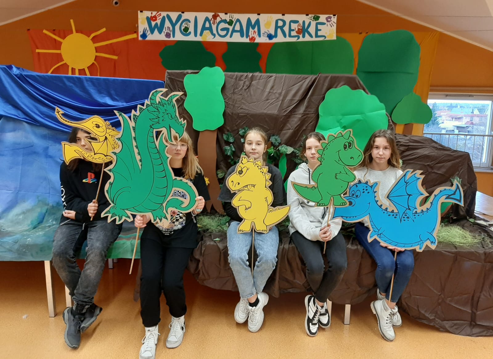 Wyciągam rękę Na obrazku widać 4 dziewczynki siedzące na scenie i trzymające smoki namalowane na kartonach