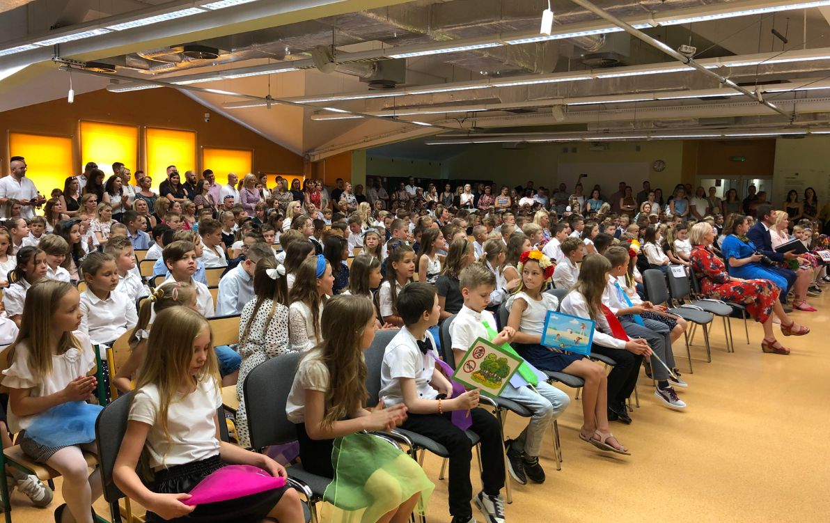 Zakończenie 2022 Na zdjęciu widać uczniów klas 1 i 2 siedzących w auli na krzesłach podczas uroczystości zakończenia roku szkolnego.