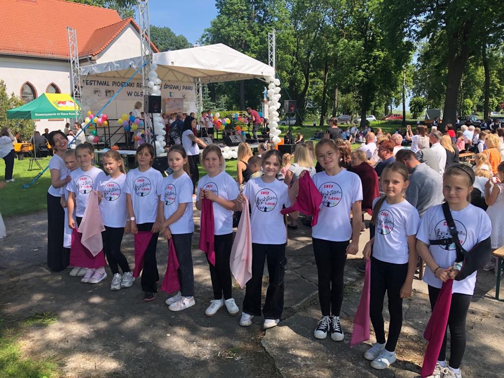 Ch&oacute;rzystki wraz z&nbsp;dyrygentką pozują ustawione w&nbsp;szeregu na&nbsp;świeżym powietrzu ubrane  w&nbsp;białe koszulki z&nbsp;logo ch&oacute;ru Bambino,, trzymają w&nbsp;dłoniach r&oacute;żowe chusty.