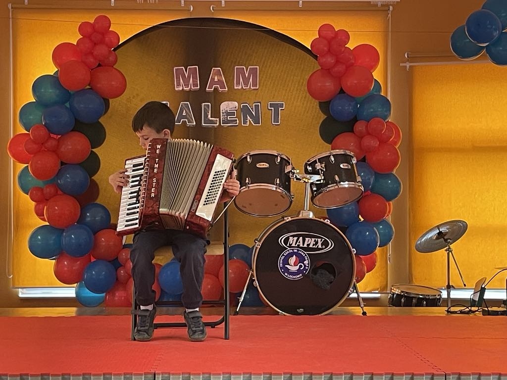 Jeden z&nbsp;uczestnik&oacute;w konkursu szkolnego "Mam talent" grający na&nbsp;akordeonie, uczeń siedzi na&nbsp;krześle na&nbsp;scenie, w&nbsp;tle perkusja oraz&nbsp;napis "Mam talent".