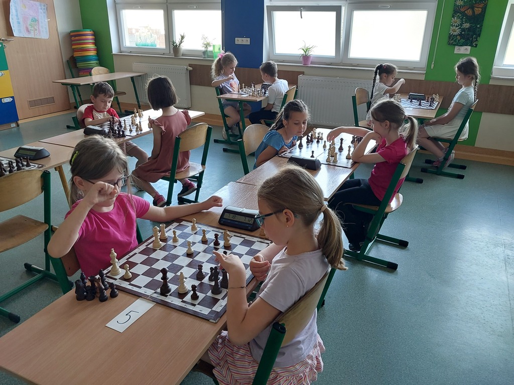 Na zdjęciu dzieci z&nbsp;oddział&oacute;w przedszkolnych podczas rozgrywek szachowych, siedzący parami na&nbsp;przeciw siebie w&nbsp;ławkach szkolnych, pomiędzy nimi plansza do szach&oacute;w.