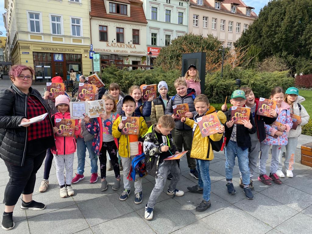 Uczniowie oddziału 1c wraz z wychowawcą pozują do zdjęcia na rynku w Bydgoszczy. Uczniowie trzymają w dłoniach książeczki zawierające zagadki/zadania do wykonania.