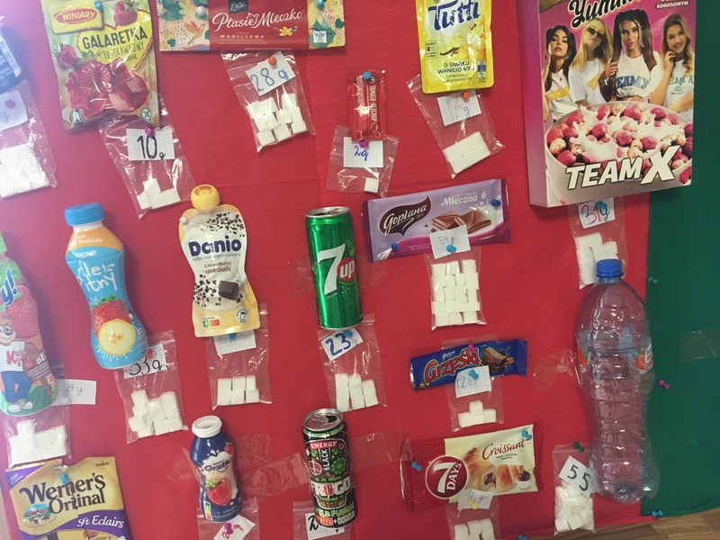 Tablica edukacyjna przedstawiająca zawartość cukru w produktach Zdjęcie przedstawia tablicę edukacyjną, na której zamieszczone są wybrane produkty oraz ilość cukru, jaką zawierają