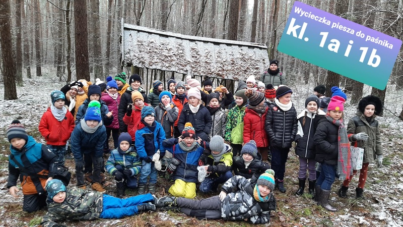 1a i 1b dokarmiają leśne zwierzęta Zdjęcie przedstawia uczniów klas 1a i 1b zimą w lesie, na tle paśnika, gdzie dostarczyli pożywienie dla zwierząt.