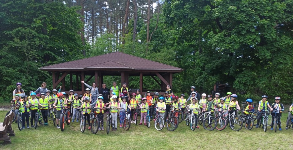 2b i 2d na rowerach w Leśnictwie Nadkanale Na pierwszym planie uczniowie na rowerach, za nimi opiekunowie, altana i las w tle.