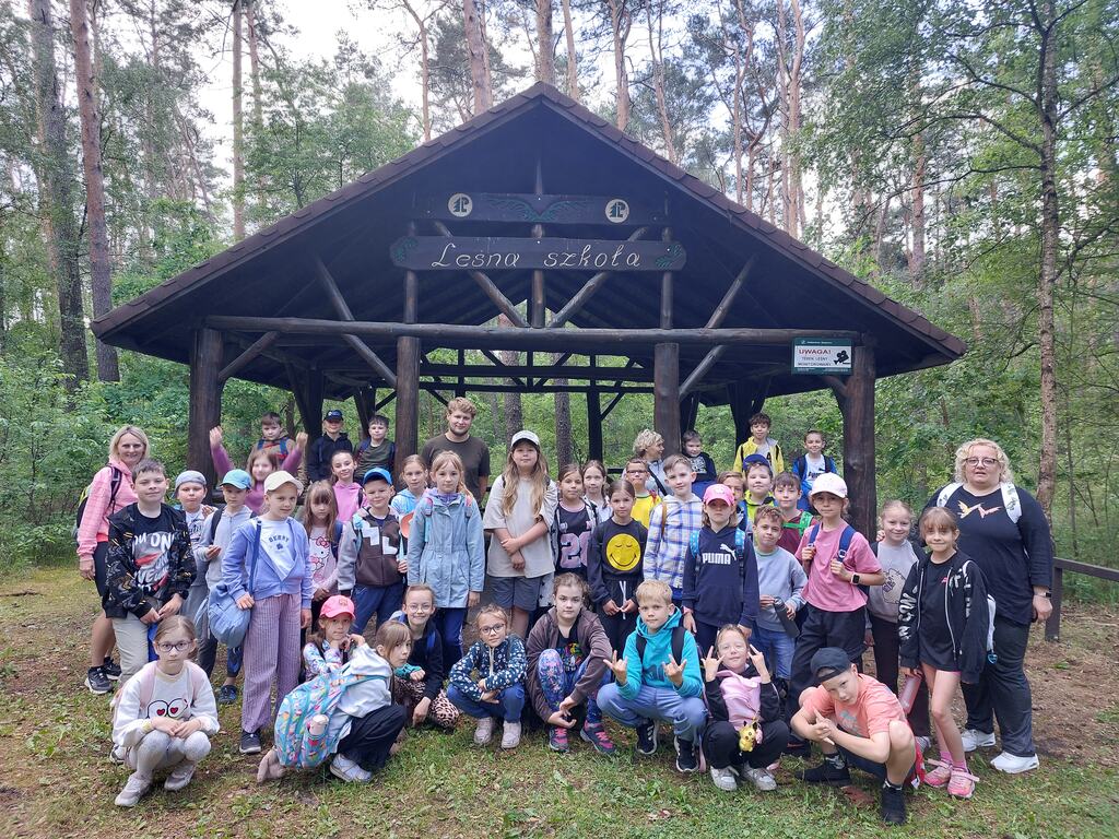 Zdjęcie przedstawia uczni&oacute;w klas 2a i&nbsp;2c wraz z&nbsp;wychowawczyniami w&nbsp;lesie przed altaną z&nbsp;napisem "Leśna szkoła"