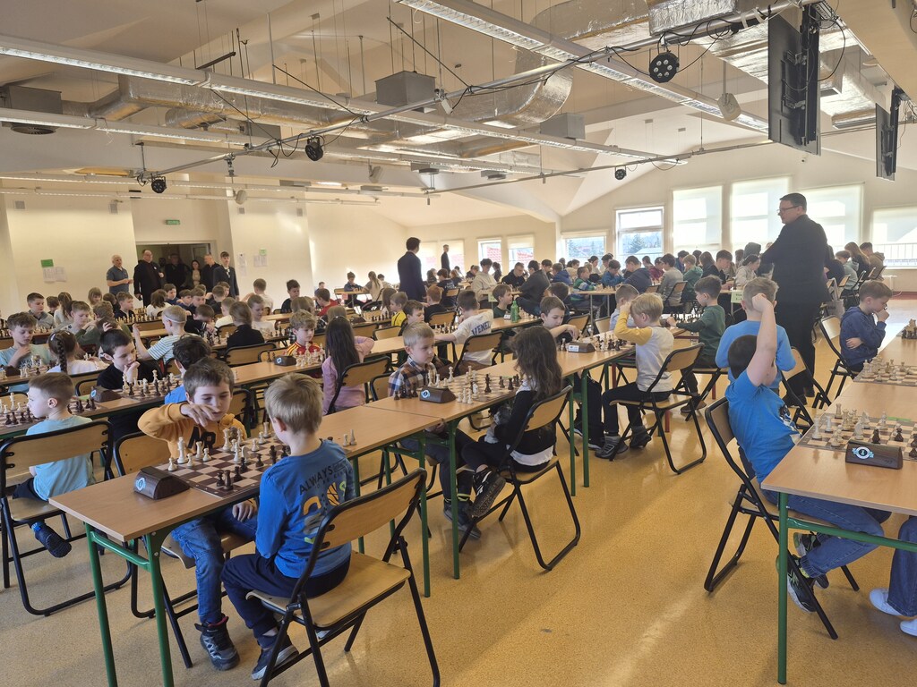 Zdjęcie ukazuje uczestnik&oacute;w turnieju szachowego zajmujących miejsce przy stolikach i&nbsp;toczących b&oacute;j pionami nad szachownicami.