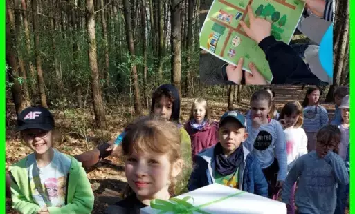 Zdjęcie przedstawia dziewczynkę z prezentem na tle dzieci z jej klasy w lesie. W prawym górnym rogu widnieje mapa, wykonana przez rodziców uczniów klasy 1.
