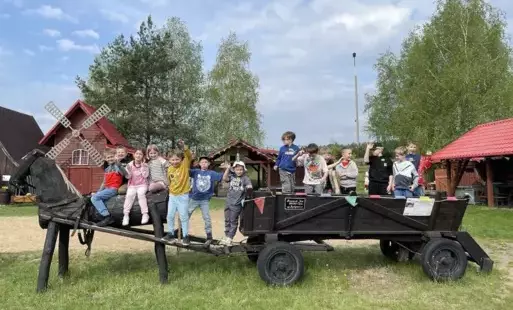 Uczniowie klas pierwszych w Janiej Górze pozujący na drewnianym wozie z koniem z drewna, w tle krajobraz wiejski.