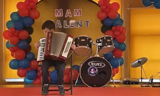 Jeden z uczestników konkursu szkolnego "Mam talent" grający na akordeonie, uczeń siedzi na krześle na scenie, w tle perkusja oraz napis "Mam talent".