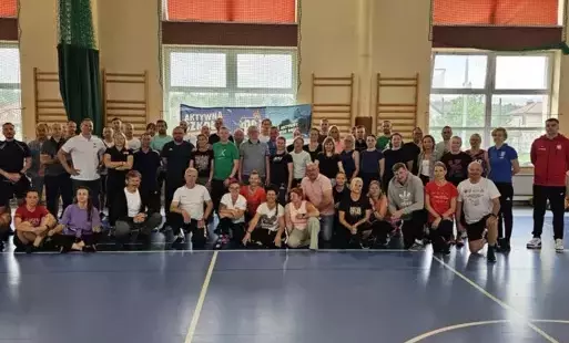 Do zdjęcia pozują nauczyciele wychowania fizycznego na sali gimnastycznej, ustawieni w zwartej grupie 64-osobowej, niektórzy stoją, inni kucają, ubrani są w stroje sportowe.