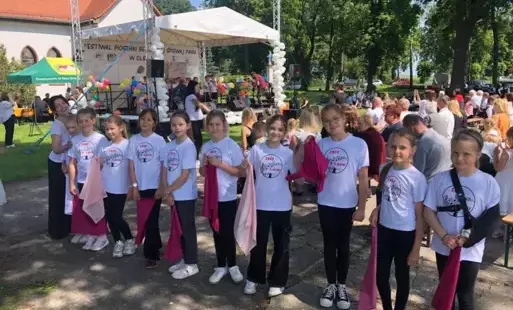 Chórzystki wraz z dyrygentką pozują ustawione w szeregu na świeżym powietrzu ubrane  w białe koszulki z logo chóru Bambino,, trzymają w dłoniach różowe chusty.