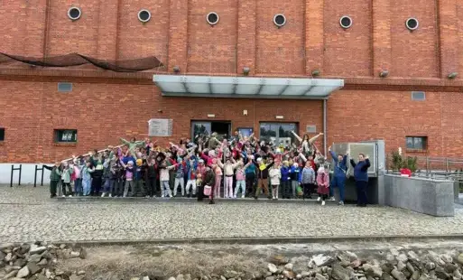 Uczniowie klas 1 SP Łochowo pozują z rękami uniesionymi do góry przed wejściem do Młynu Wiedzy w Toruniu.