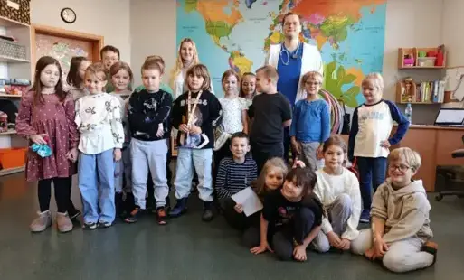 Do zdjęcia pozuje grupa stojących dzieci, za nimi doktor w białym kitlu oraz pani, w tle mapa świata.