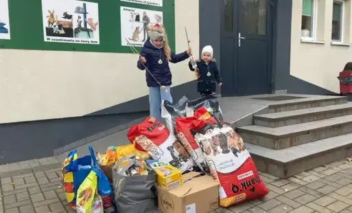 Na zdjęciu widać dwójkę uczniów, którzy stoją przed schroniskiem. Przed nimi zebrane materiały i pokarm dla czworonogów ze schroniska.