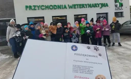 Uczniowie klasy 2a wraz z wychowawczynią pozują do zdjęcia z pluszakami. Za nimi przychodnia weterynaryjna. Na pierwszym planie wydrukowane podziękowanie dla przychodni. Sceneria zimowa.