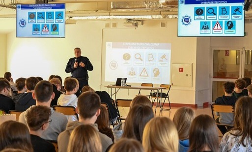 Na zdjęciu widać policjanta, który z użyciem prezentacji multimedialnej prowadzi szkolenie dotyczące cyberprzemocy w auli szkolnej dla uczniów z klas 4-8. Widać również siedzących tyłem uczniów, skierowanych w stronę prelegenta.