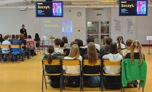 Zdjęcie przedstawia uczniów siedzących na auli i słuchających wykładu prowadzonego przez Mr. Universe w kulturystyce Tomasza Gizę na auli szkolnej.