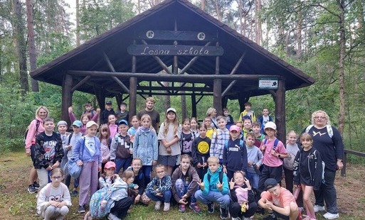 Zdjęcie przedstawia uczniów klas 2a i 2c wraz z wychowawczyniami w lesie przed altaną z napisem "Leśna szkoła"